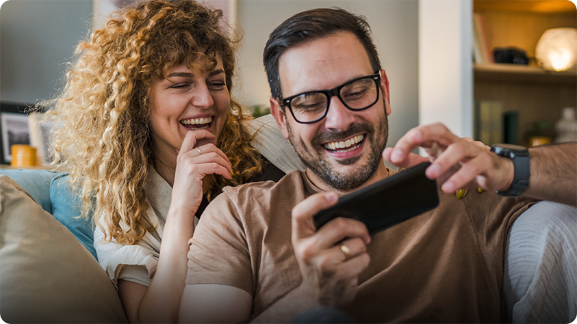 Zwei Personen sitzen auf einem Sofa und schauen gemeinsam auf ein Smartphone, mit Regalen und Wohnaccessoires im Hintergrund.