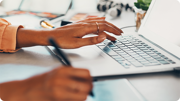 Nahaufnahme von Händen bei der Arbeit an einem Schreibtisch – eine Hand tippt auf einem Laptop, die andere hält einen Stift über einem Notizblock, mit unscharfen Bürogegenständen im Hintergrund.