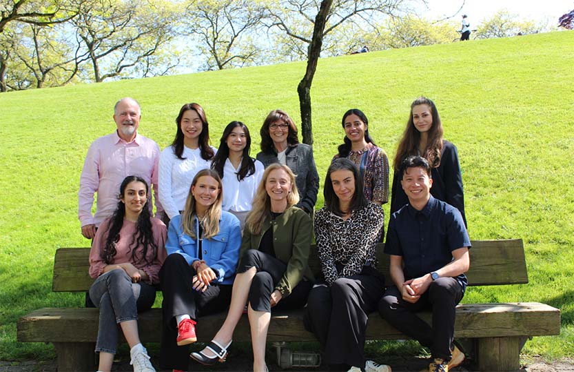 A group of ten people are posing for a photo outdoors on a sunny day. They are sitting and standing on and around a wooden bench in a park with green grass and trees in the background.