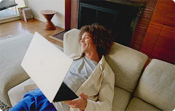 A person is lying on a beige couch in a living room, holding an open laptop.