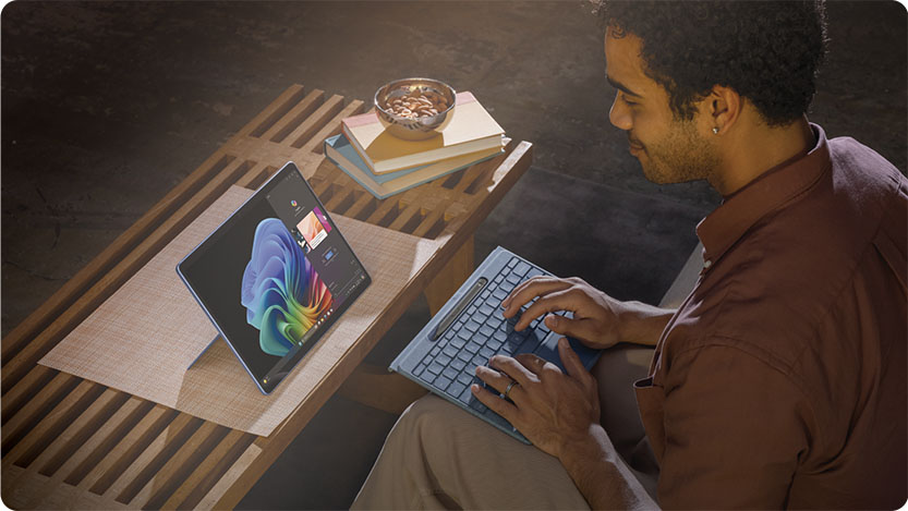 A person sits on a sofa, using a tablet with a detachable keyboard on their lap. The tablet is propped up on a coffee table, displaying colorful graphics.