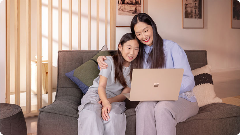 Two people sitting on a gray couch, one holding a laptop. The person on the left is leaning in close, and both appear to be looking at the laptop screen.