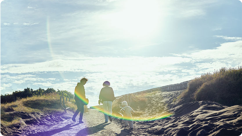 A photograph of two adults and a small child walking along a sandy path surrounded by grassy dunes under a bright sky.