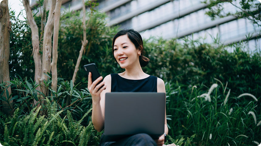 A person sitting outdoors in a green park-like setting, holding a smartphone in one hand and a laptop on the lap.