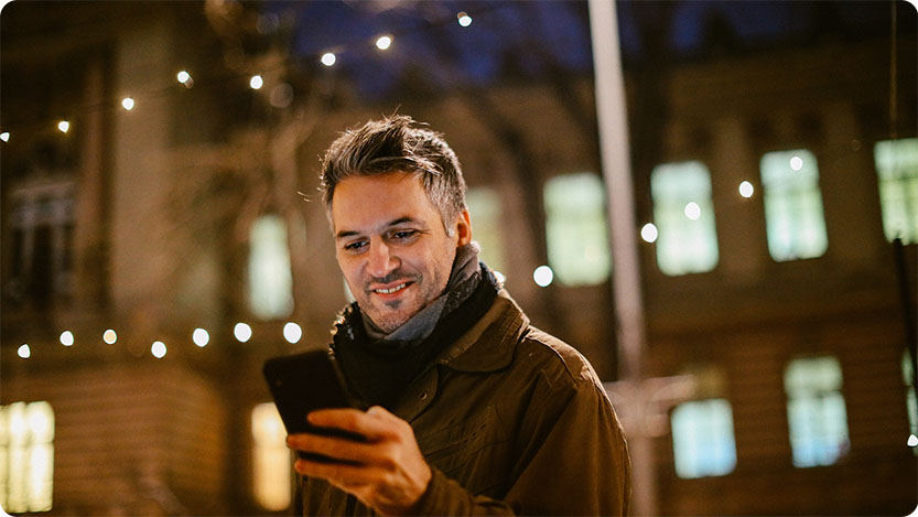 Person outdoors at night holding a phone, with string lights and lit windows in the background.