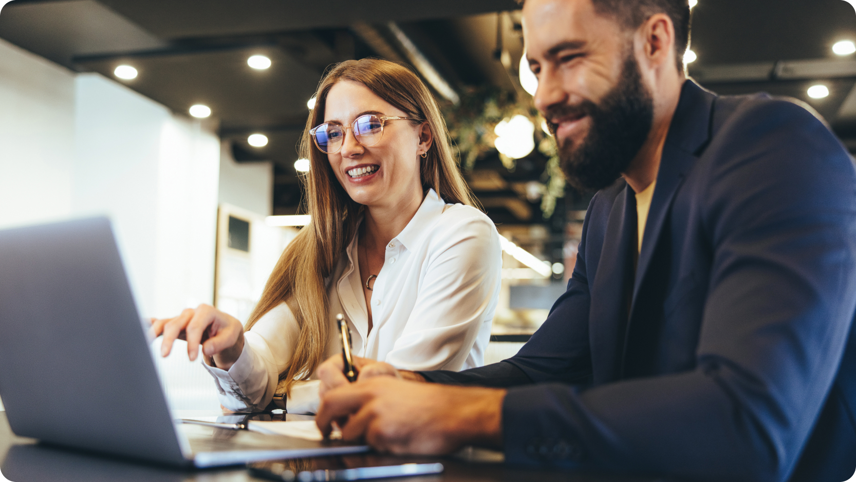 Two professionals collaborating at a laptop in a modern office setting, reviewing documents and discussing strategy