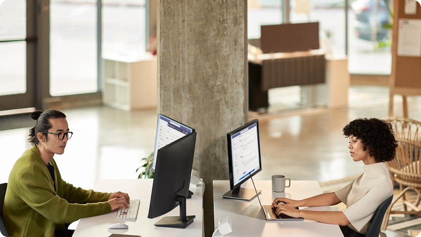 Two people work quietly across from each other at a shared desk in a bright, modern office, each focused on their own computer in an open, calm workspace.