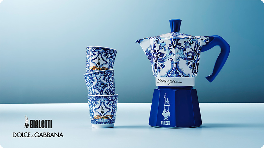 A blue and white patterned Bialetti moka pot from the Dolce & Gabbana Blu Mediterraneo collection, placed on a light surface against a gradient blue background. Next to it, three matching espresso cups are stacked vertically. The Bialetti and Dolce & Gabbana logos appear in the bottom left corner.