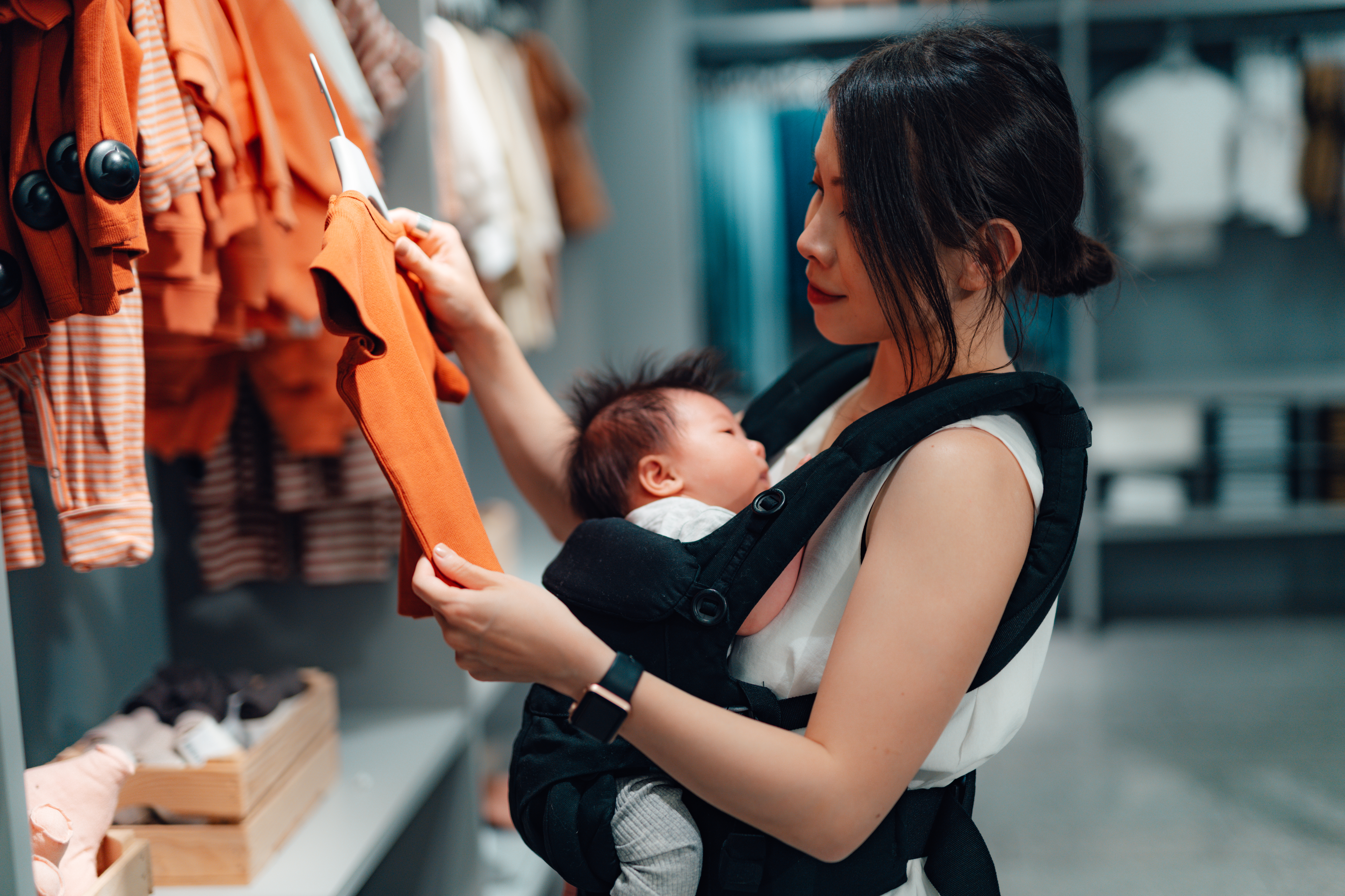 Retail shopper selecting baby clothes in a children’s clothing store while carrying an infant