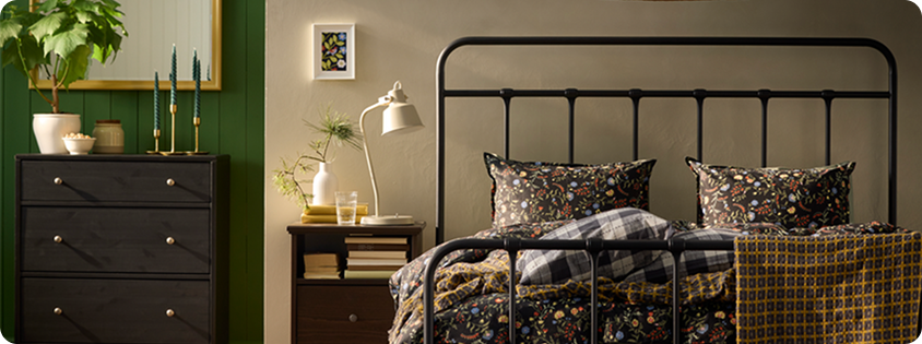 Cozy IKEA bedroom with a black metal bed, patterned bedding, dark wood furniture, and green panelled walls