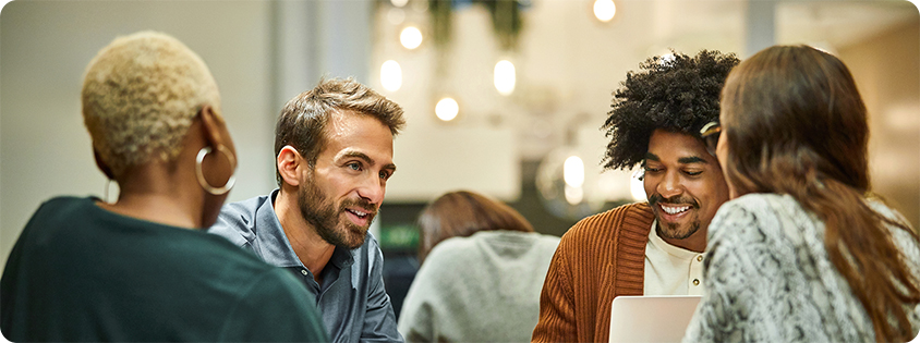 Group of coworkers sitting around a table, collaborating over a laptop and printed materials in a modern office setting.