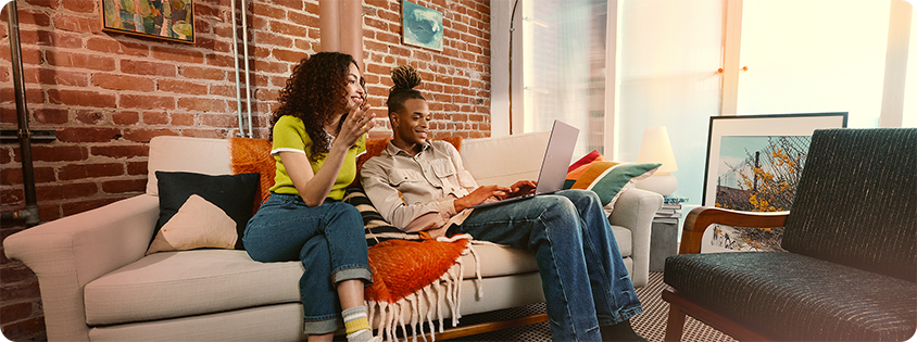 Two people sitting on a sofa using a laptop in a modern living room with exposed brick walls and natural light.